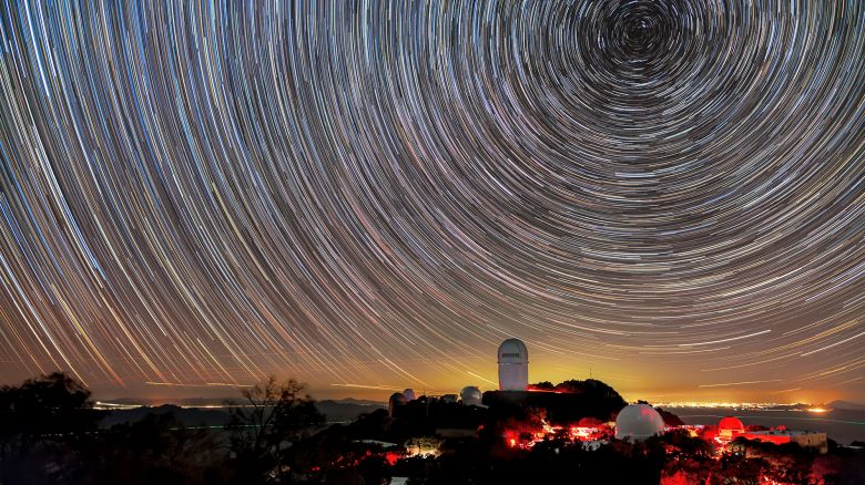 These whirling lines in the sky are the trails of stars after an hour-long exposure above Kitt Peak National Observatory (KPNO), a Program of NSF NOIRLab. The trails are shortest around the North Star, Polaris, a star that happens to coincide almost directly with the celestial north pole. The different colors in the trails reflect the different temperatures of the stars, with blue being the hottest stars and yellow/red the coolest. The telescope visible above the horizon is the Nicholas U. Mayall 4-meter Telescope, and the red glow on the mountain is caused by red lights used to ensure the eyes of visitors and staff remain dark adapted at night. The yellow glow on the horizon comes from Tucson, AZ, about 89 kilometers (55 miles) to the northeast. Over the past decade, light pollution like this has increased by 2% a year from many medium-sized and larger cities. To help monitor light pollution in your city, you can participate in the citizen science campaign, Globe at Night.
